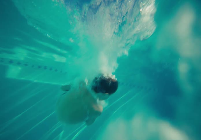 A young boy cannonballs in slow motion off a diving board into a backyard swimming pool on a bright sunny day