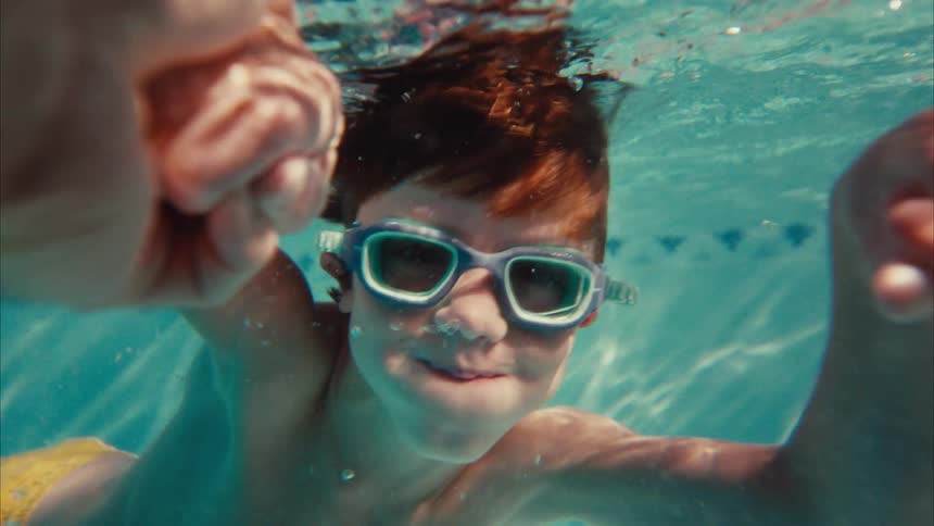 A smiling red-headed boy wearing goggles playfully swims towards the camera underwater in a swimming pool