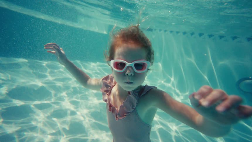 Young girl wearing pink swim goggles glides underwater in a sunlit swimming pool
