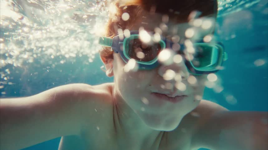 Underwater selfie point of view of a young boy with red hair wearing goggles and swimming in a sunlit pool