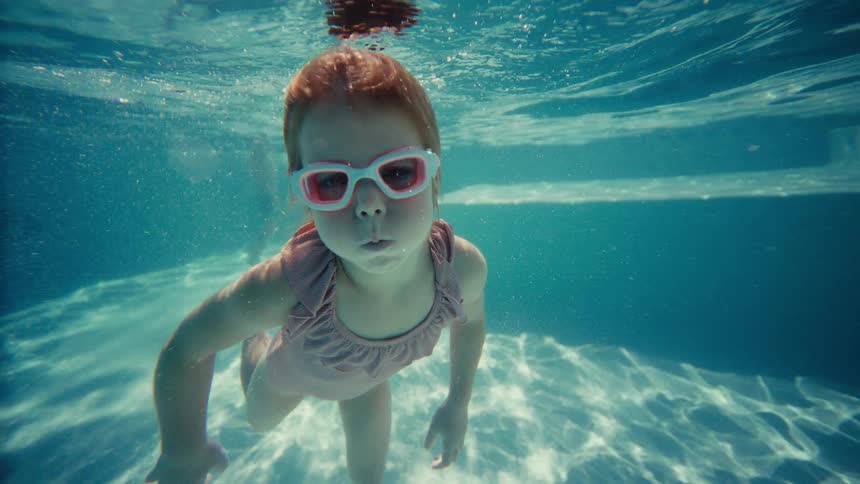 Little girl in pink goggles swims underwater in a sunlit pool