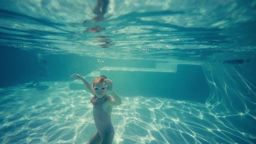 Young girl wearing swim goggles plays underwater in a sunlit swimming pool