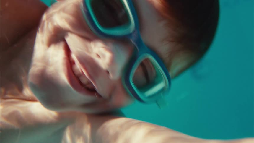 A happy young boy wearing goggles smiles and floats on his back just beneath the surface of the water in a pool, shot in slow motion