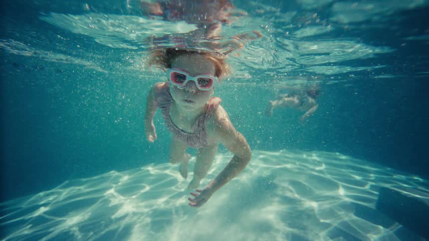 Underwater slow-motion view of a young girl in goggles swimming gracefully towards the camera in a sunlit pool