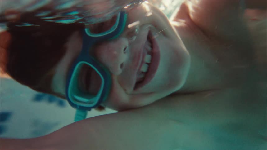 Slow motion underwater close-up of a happy boy with red hair and goggles smiling in a swimming pool
