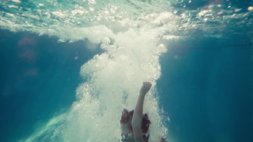 A smiling boy in goggles cannonballs into a swimming pool, captured in a slow-motion underwater shot with bubbles all around