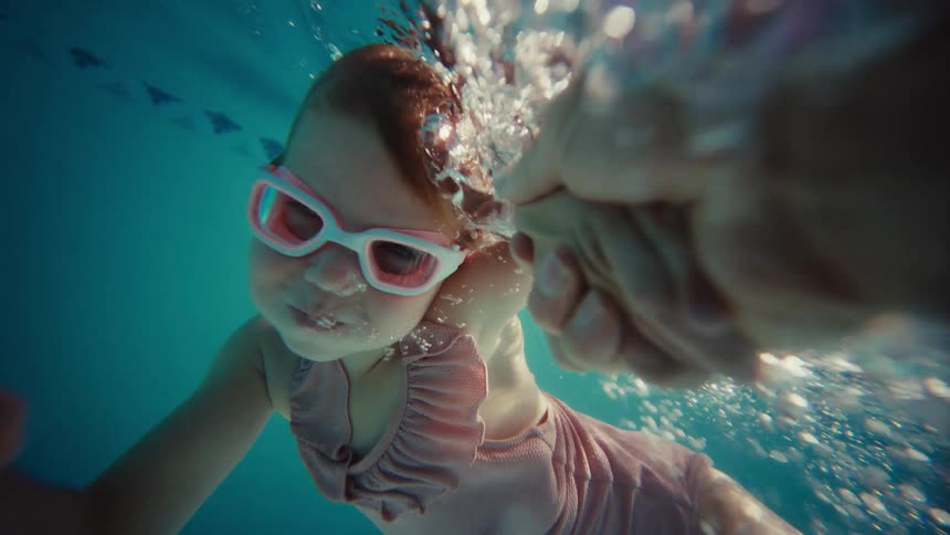 A father holds his young daughter's hand and guides her as she swims underwater in slow motion