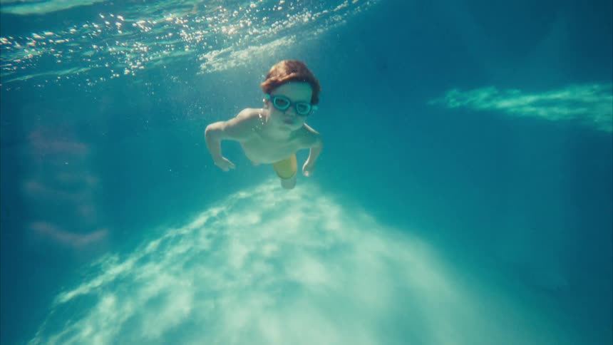 Slow motion underwater view of a young boy with goggles swimming gracefully toward the camera in a sunlit pool