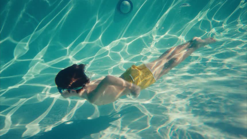 A young boy in yellow swim trunks gracefully glides underwater in a sunlit swimming pool in slow motion