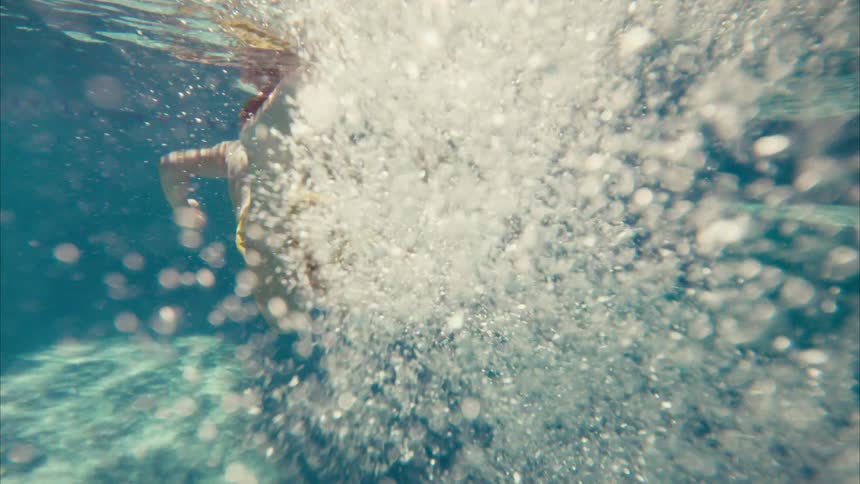 An underwater slow-motion view shows a young boy in yellow shorts swimming away, kicking his feet and creating a stream of bubbles in a sunlit pool