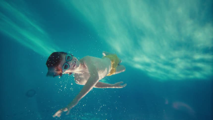 Slow motion underwater shot of a cheerful boy in goggles floating and swimming in a bright blue pool