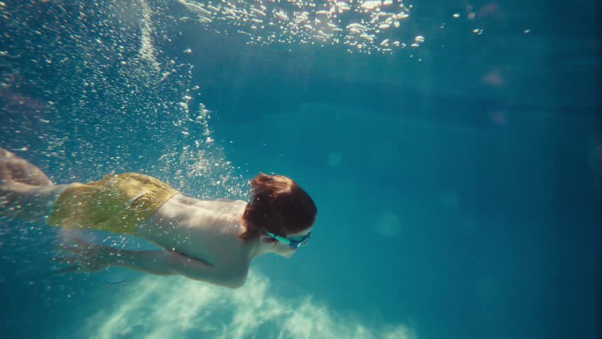 An underwater slow-motion shot captures a young boy with red hair and goggles swimming gracefully in a sunlit pool