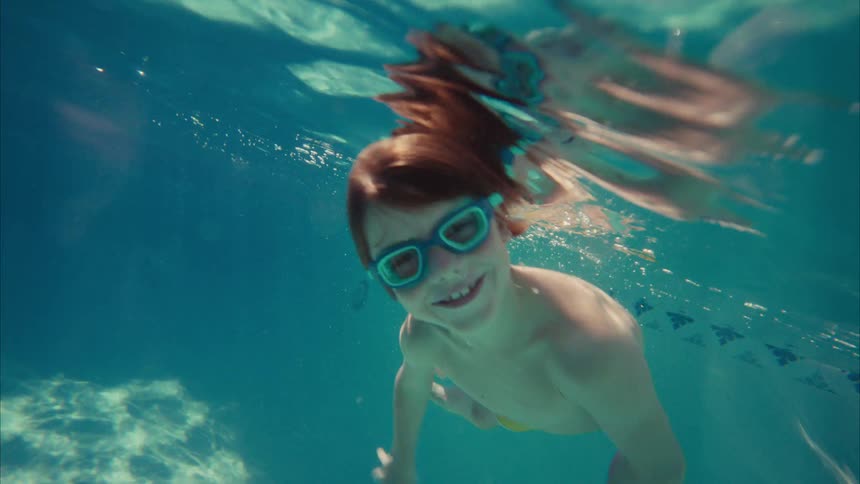 A smiling red-haired boy wearing goggles swims towards the camera underwater in a sunlit swimming pool in slow motion