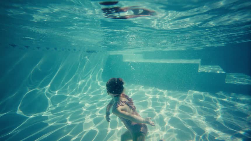 Young girl in swim goggles playfully dancing underwater in a sunlit swimming pool