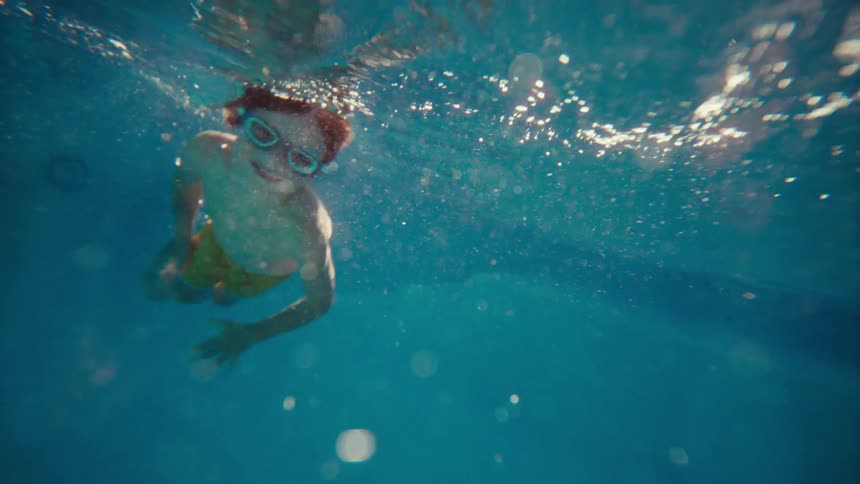 A happy young boy with red hair and goggles smiles as he swims underwater in a sunlit pool