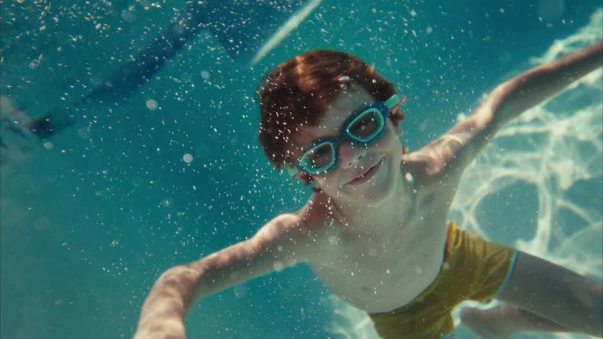 A happy boy with red hair and goggles smiles while gracefully swimming underwater in a sunny pool