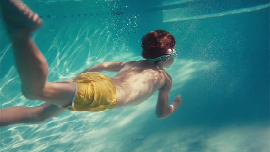 A young boy with red hair swims underwater in a sunlit swimming pool in slow motion