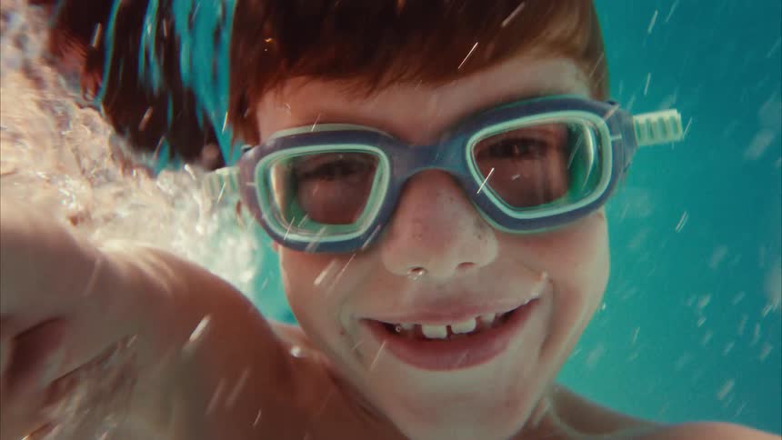 A happy boy with red hair and swimming goggles smiles wide for the camera in slow motion while underwater in a pool