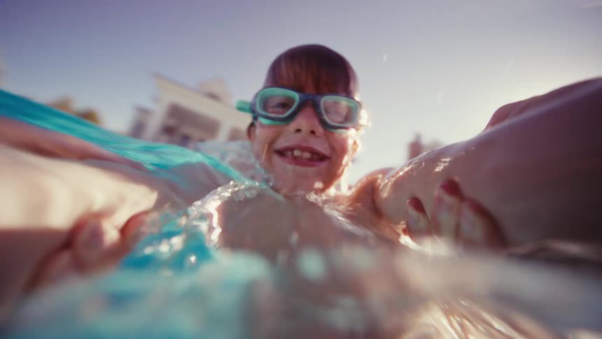 Smiling boy with goggles takes a playful selfie video while swimming and emerging from the water in a sunlit pool
