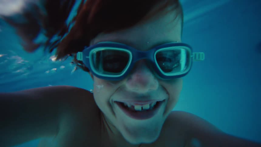 An adorable boy wearing swimming goggles smiles wide for a selfie while floating underwater in a bright blue pool in slow motion