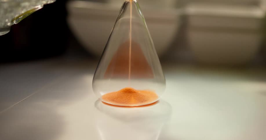 A close-up shot shows orange sand falling through the narrow neck of an hourglass and piling up at the bottom
