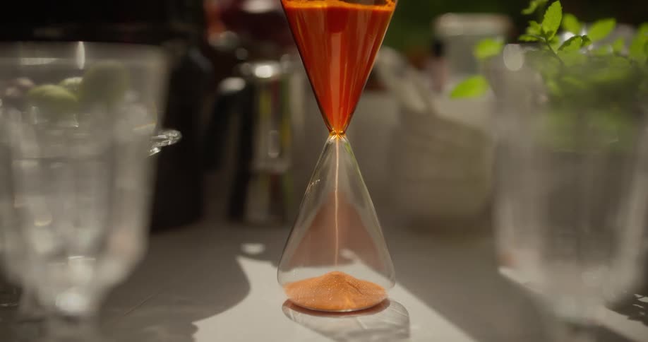 A modern hourglass with orange sand counts down the time as the camera slowly tilts down in a sunlit setting