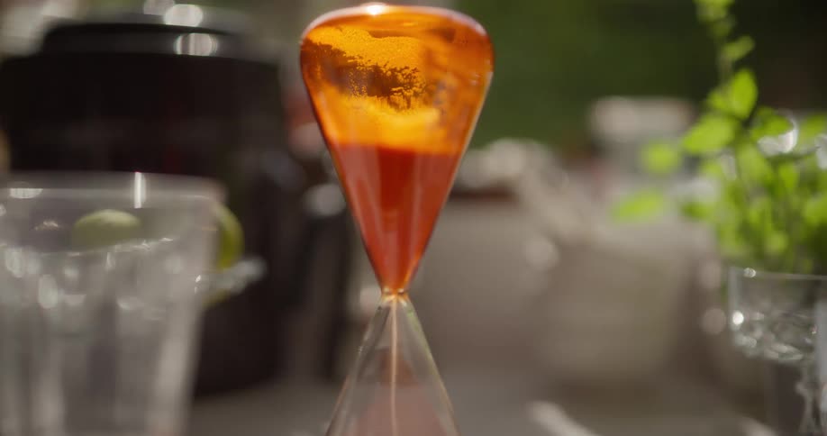 Slow dolly shot focuses on a modern orange hourglass with sand falling on a sunlit table outdoors