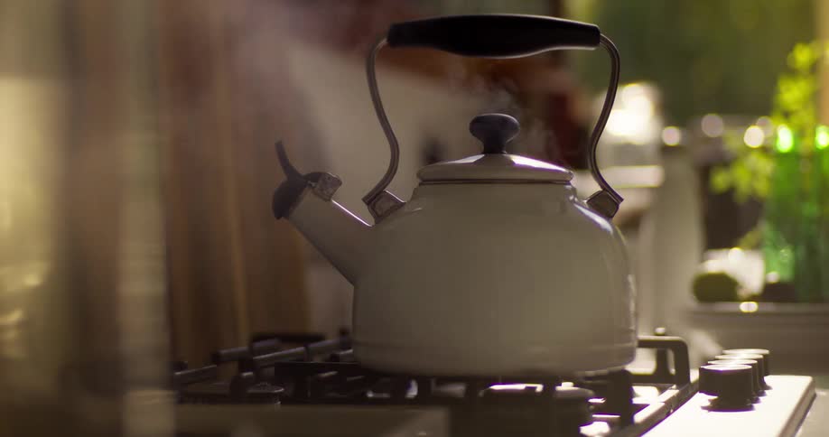 Steam rises from a white kettle as a hand turns on the gas stove to boil water