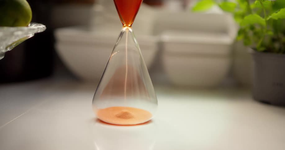 A close-up of a modern glass hourglass with orange sand measuring the passing of time on a white surface