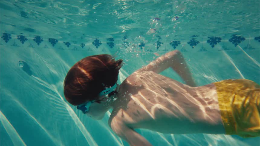 A young boy wearing goggles swims underwater in a sunlit pool in slow motion