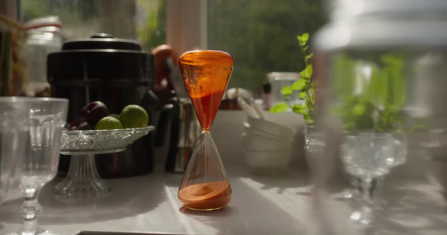 A bright orange hourglass measures the passage of time as sand falls on a sunlit kitchen counter