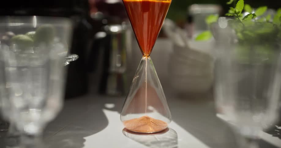 An elegant, modern hourglass with orange sand measures the passage of time on a sunlit tabletop