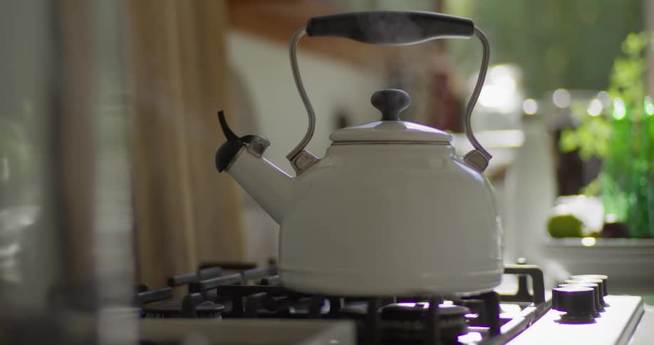A hand turns on the gas stove to boil water in a classic white kettle, which begins to release steam in a sunlit kitchen