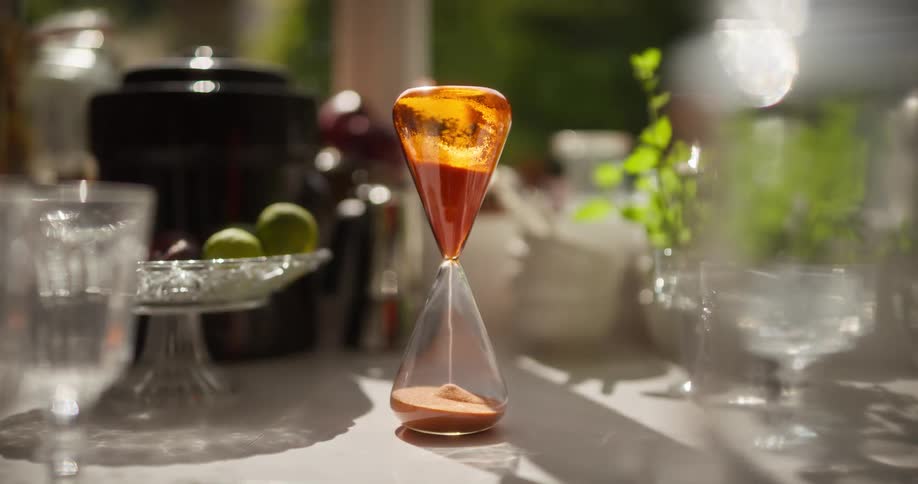 A rack focus reveals a stylish orange hourglass measuring time on a beautifully set table in the sun