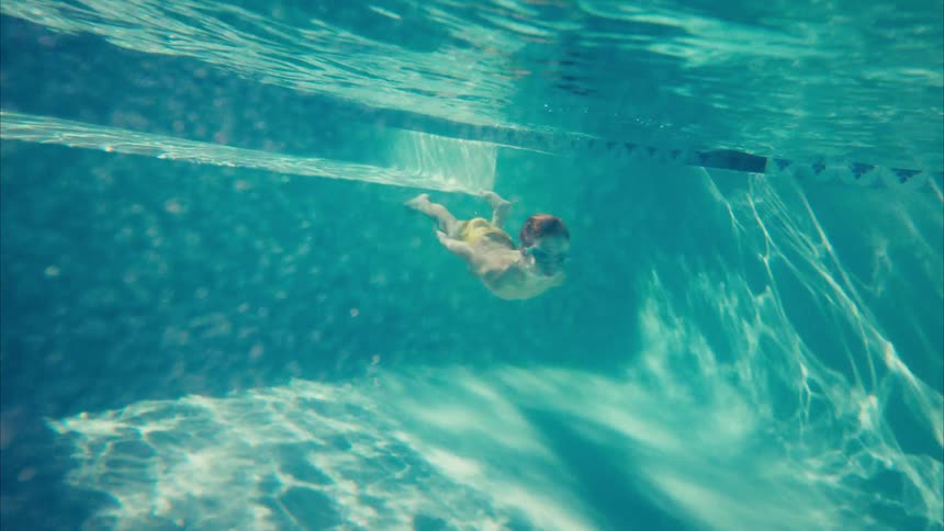 A young boy with goggles gracefully swims underwater in a sunlit swimming pool in slow motion