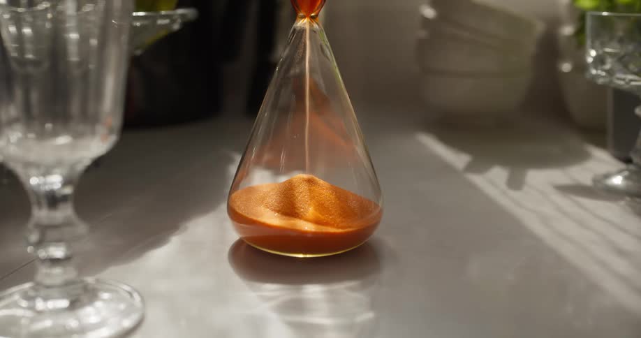 Golden sand trickles through a glass hourglass, measuring time on a sunlit kitchen countertop