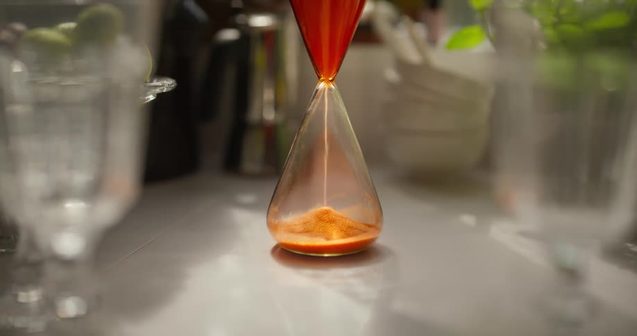 Orange sand trickles through a glass hourglass, measuring the passage of time on a sunlit kitchen counter