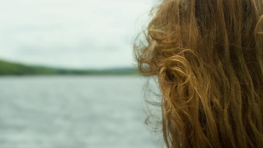 Close up from behind of a woman with curly hair blowing in the wind as she looks out over a lake in slow motion