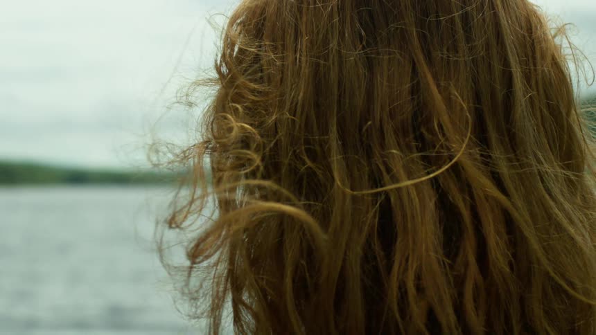 Slow-motion close-up of a woman's hair blowing in the wind as she gazes thoughtfully across a peaceful lake