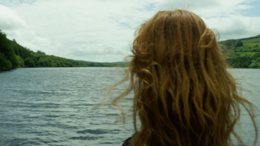 An over-the-shoulder shot captures a woman with long red hair as she peacefully contemplates a serene lake, with the focus shifting from the landscape to her hair blowing in the wind