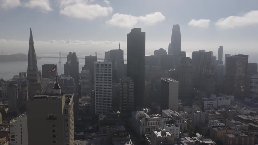 A slow aerial view pulls back from the iconic San Francisco skyline, revealing the Transamerica Pyramid and Bay Bridge on a hazy day