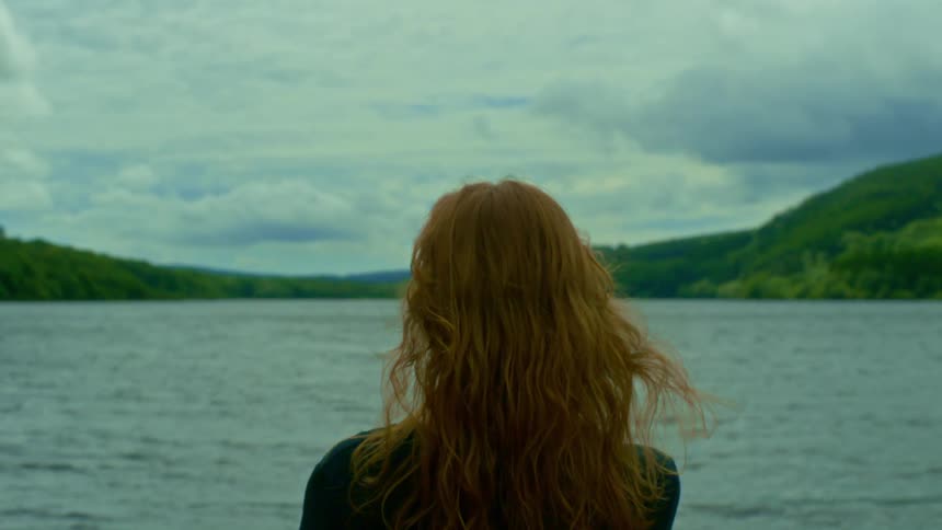 A redhead stands peacefully, watching a serene lake as the wind gently blows through her hair