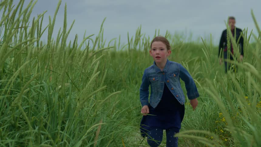 A young girl runs in slow motion through a field of tall green grass as her father walks behind her