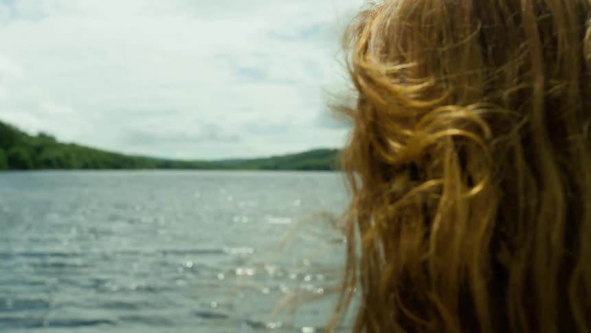 Slow motion shot of a woman's long hair blowing in the breeze as she looks out over a beautiful, sunlit lake
