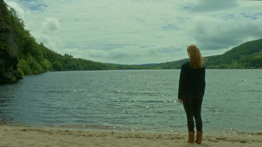 A woman stands alone on a sandy shore, her hair blowing in the wind as she gazes across a tranquil lake towards distant forested hills