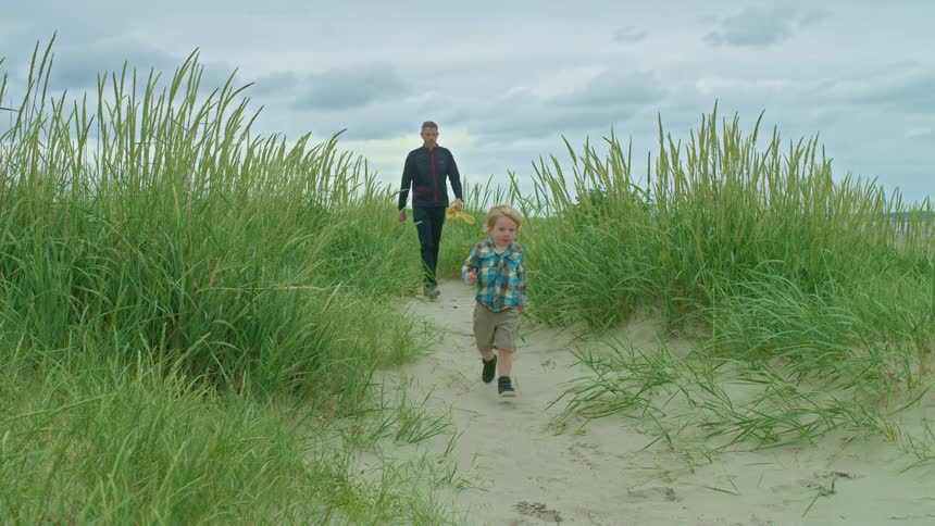 A young boy runs in slow motion along a sandy path through grassy dunes while his father walks behind him