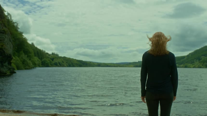 In slow motion, a woman stands contemplatively on the shore of a vast lake surrounded by green hills, her hair blowing in the wind