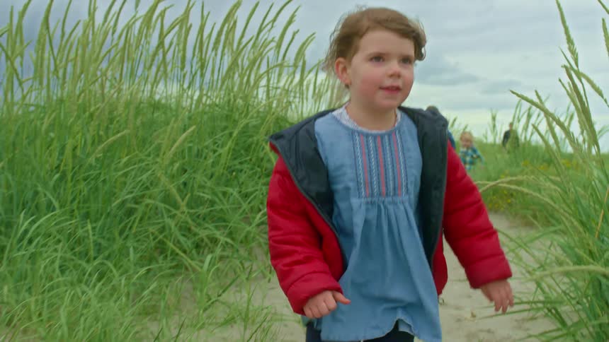 A young girl in a red jacket runs in slow motion along a sandy path through tall beach grass on a cloudy day