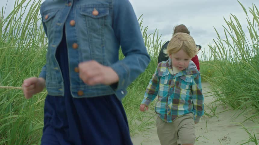 Father and his three young children happily walk in a line down a sandy path through tall beach grass in slow motion
