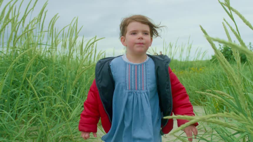 A joyful young girl in a red coat runs in slow motion along a sandy path through tall green grass, with her older sister following behind